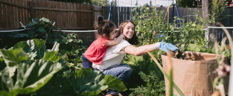 A woman pulls weeds in the garden while a child leans on her back