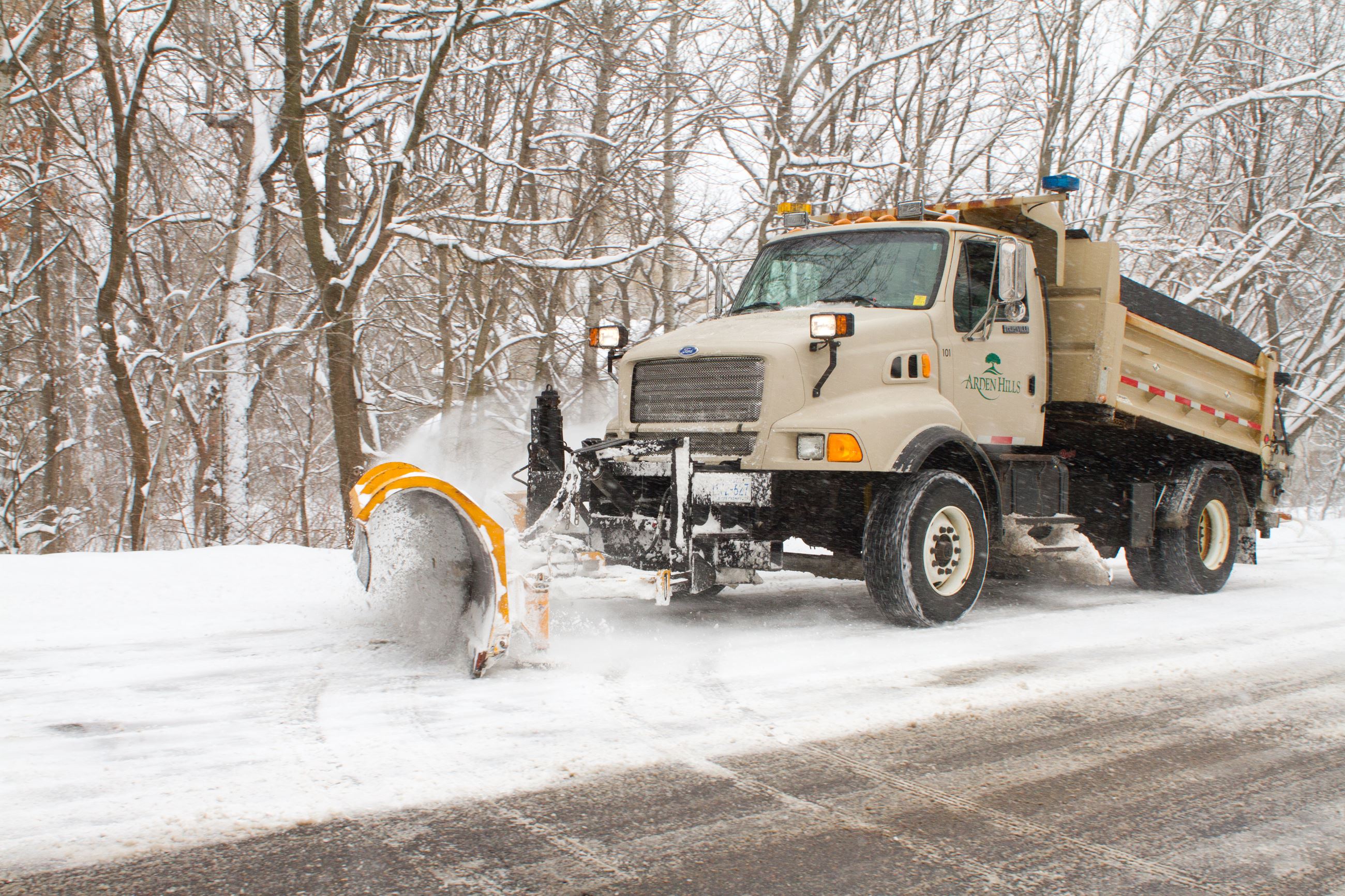 A beige snow plow plows the road with a backdrop of the snowy woods
