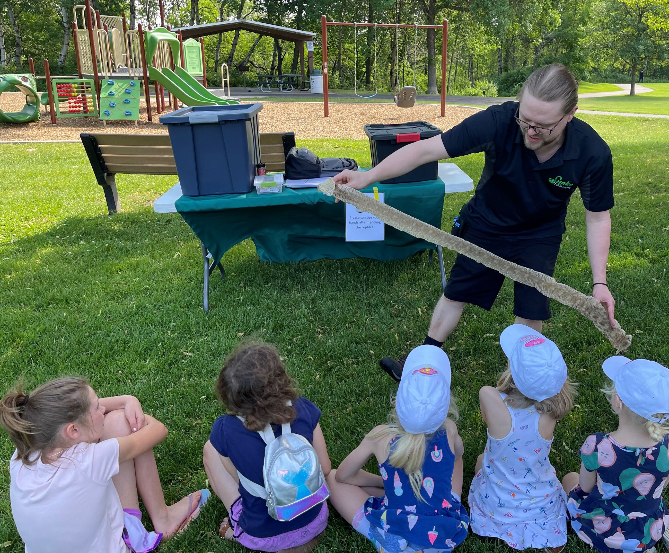 Children gather in a semi circle in front of a man who is showing them snake skins in Hazelnut park